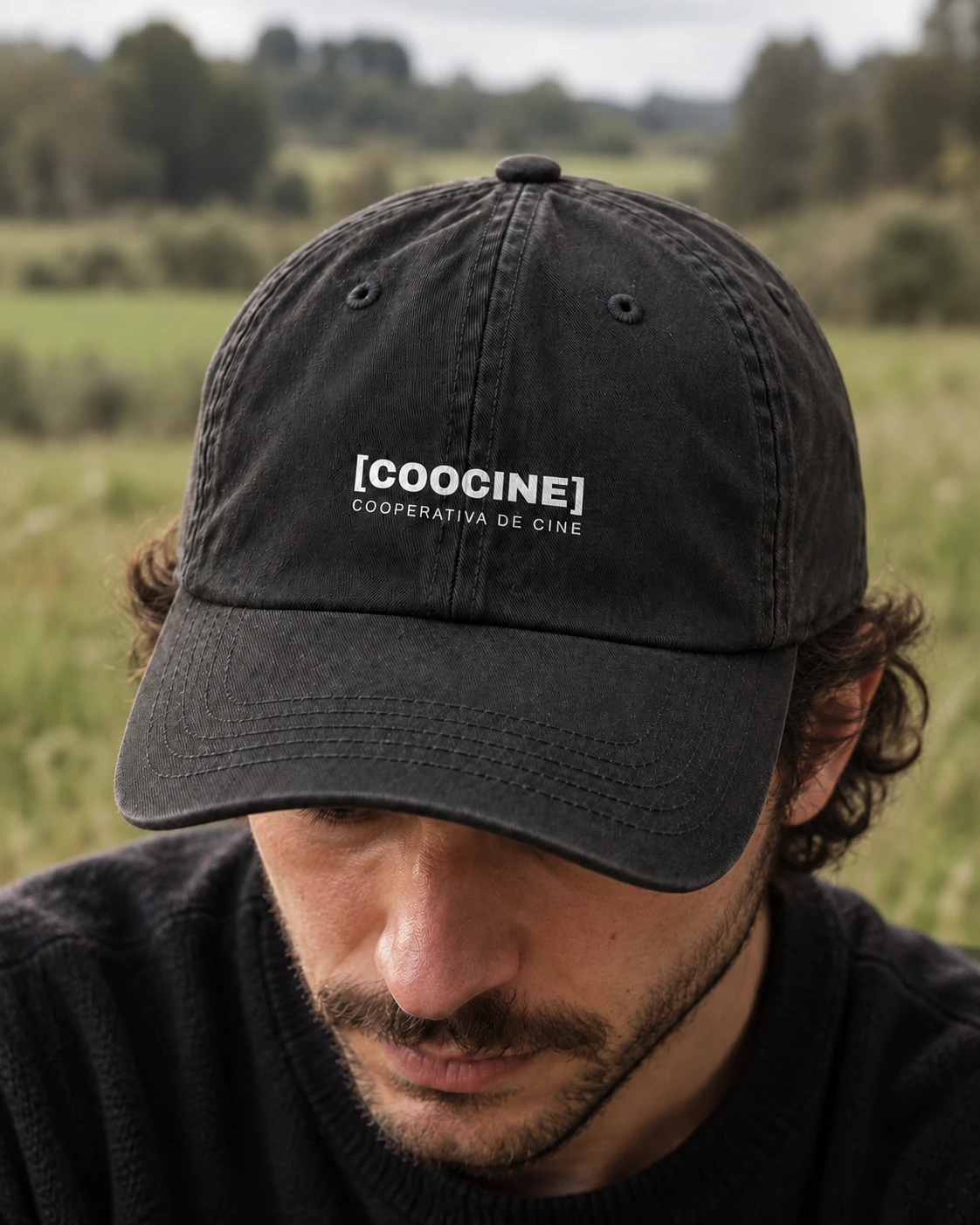 Mockup editorial de la gorra negra de Coocine puesta al aire libre, con el logo blanco pequeño centrado y el campo desenfocado al fondo.