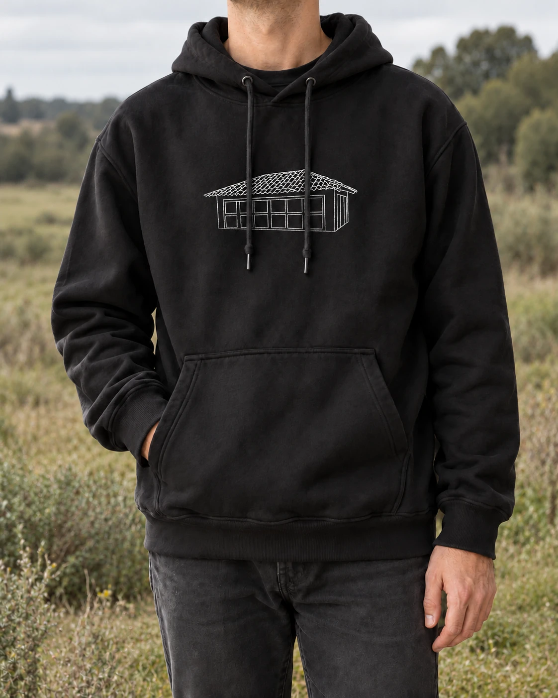 Mockup editorial del hoodie negro de Coocine puesto al aire libre, con el dibujo original de la casita en el pecho, los cordones por delante y el campo desenfocado al fondo.