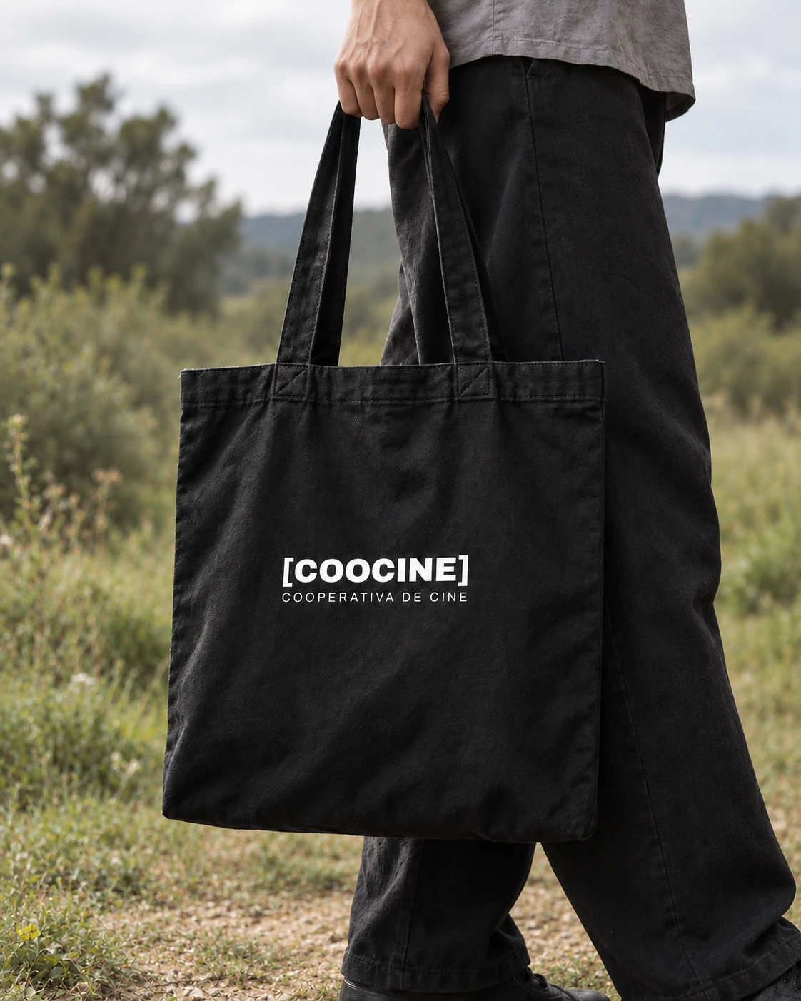 Mockup editorial de la tote negra de Coocine llevada en la mano al aire libre, con el logo blanco centrado en la bolsa y el campo desenfocado al fondo.